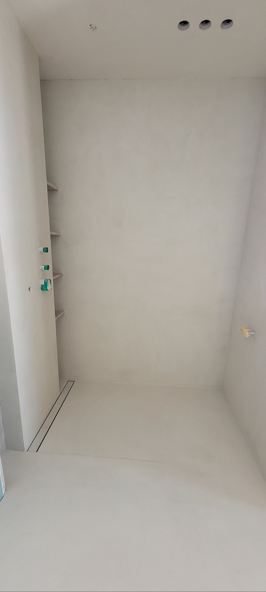 Wykończona łazienka w minimalistycznym stylu z szarym mikrocementem na ścianach i podłodze. Widoczny odpływ liniowy, półki wnękowe i punkty oświetleniowe.