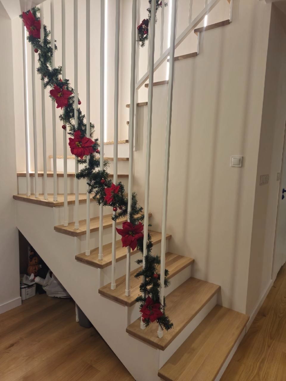 Jasne schody z drewnianymi stopniami i białymi balustradami, udekorowane świąteczną girlandą z czerwonymi kwiatami, widziane z dołu.