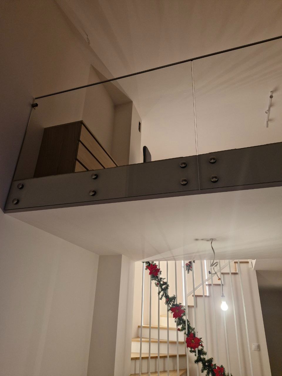 Szklana balustrada na antresoli z metalowymi mocowaniami, widok z dołu na drewniane schody z dekoracją świąteczną. Minimalistyczny design.