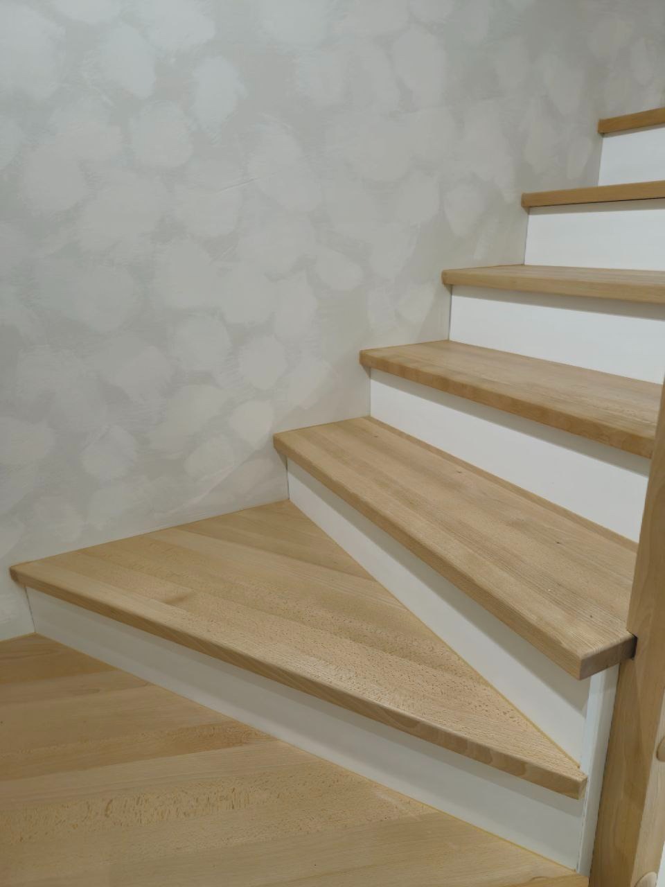 Jasne, drewniane schody z białymi podstopnicami, widoczne z dołu, z delikatną fakturą ściany w tle. Naturalne oświetlenie podkreśla detale drewna.