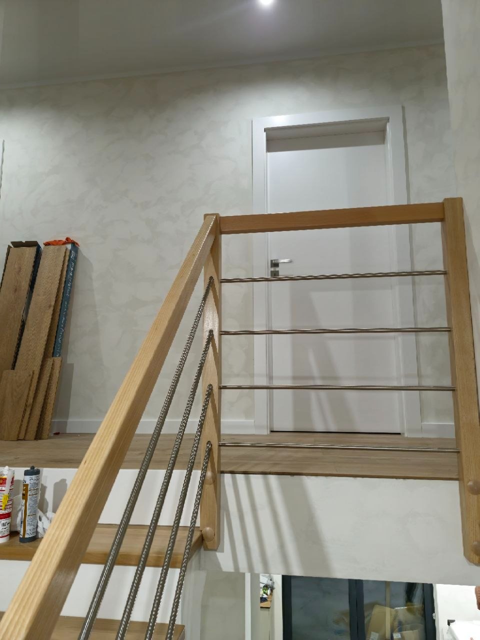 Jasne schody z drewnianą balustradą i stalowymi linkami, widok z dołu. Na ścianie widoczne płyty podłogowe i białe drzwi w tle. Minimalistyczny design.