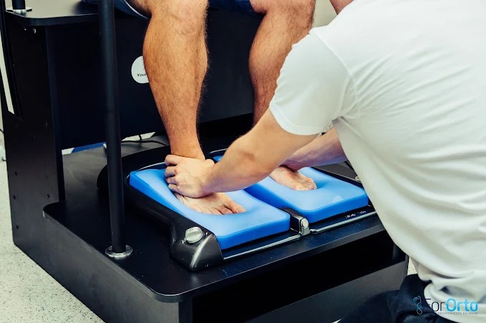 Skan stóp pacjenta na platformie diagnostycznej. Rehabilitant przykłada dłonie do stóp, przygotowując do badania. Nowoczesny sprzęt w gabinecie.