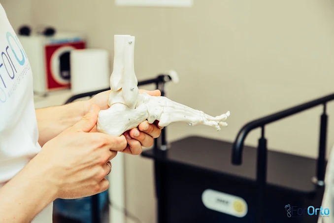 Model anatomiczny stopy w dłoniach, prezentowany w gabinecie. Widoczny fragment urządzenia rehabilitacyjnego w tle. Jasne, stonowane kolory.