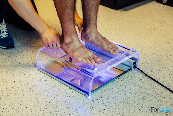 Pacjent podczas badania stóp na podoskopie z podświetleniem LED, asystent pomaga w ustawieniu. Diagnostyka wad postawy w gabinecie rehabilitacji.