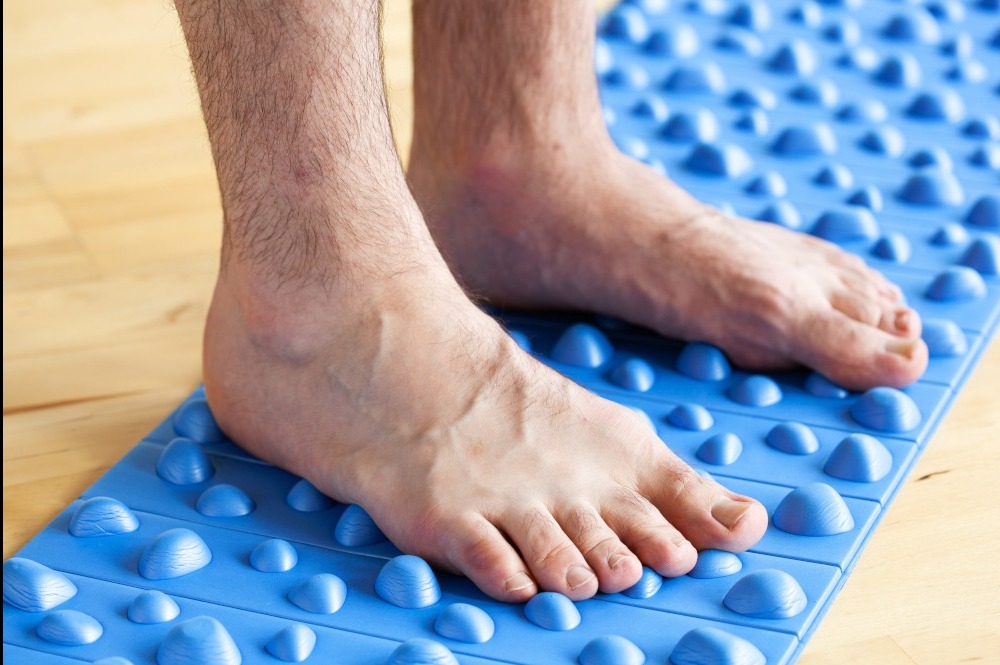 Stopy mężczyzny na niebieskiej macie do akupresury z wypustkami, na drewnianej podłodze. Bliski kadr, widoczne żyły na stopach.