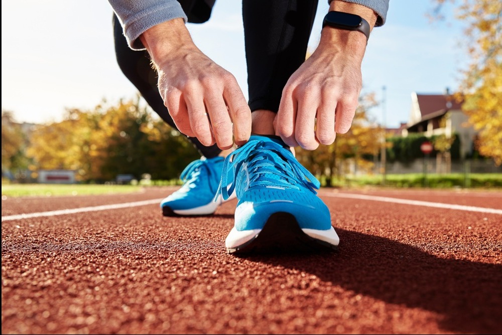 Osoba sznuruje błękitne buty sportowe na bieżni tartanowej. Widoczny zegarek sportowy na nadgarstku, jesienne drzewa w tle. Przygotowanie do treningu biegowego.