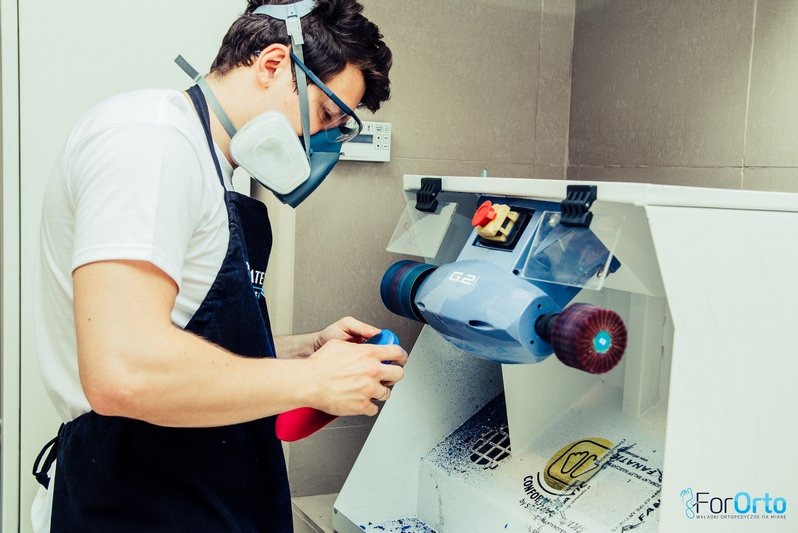 Pracownik w masce szlifuje element ortopedyczny na specjalistycznej maszynie. Widoczne drobinki materiału i logo firmy ForOrto.