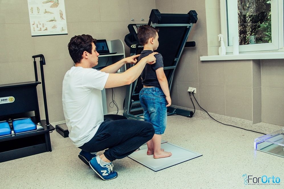 Badanie postawy dziecka przez specjalistę w gabinecie rehabilitacyjnym. Lekarz ocenia ułożenie kręgosłupa, wykorzystując platformę diagnostyczną i sprzęt rehabilitacyjny.
