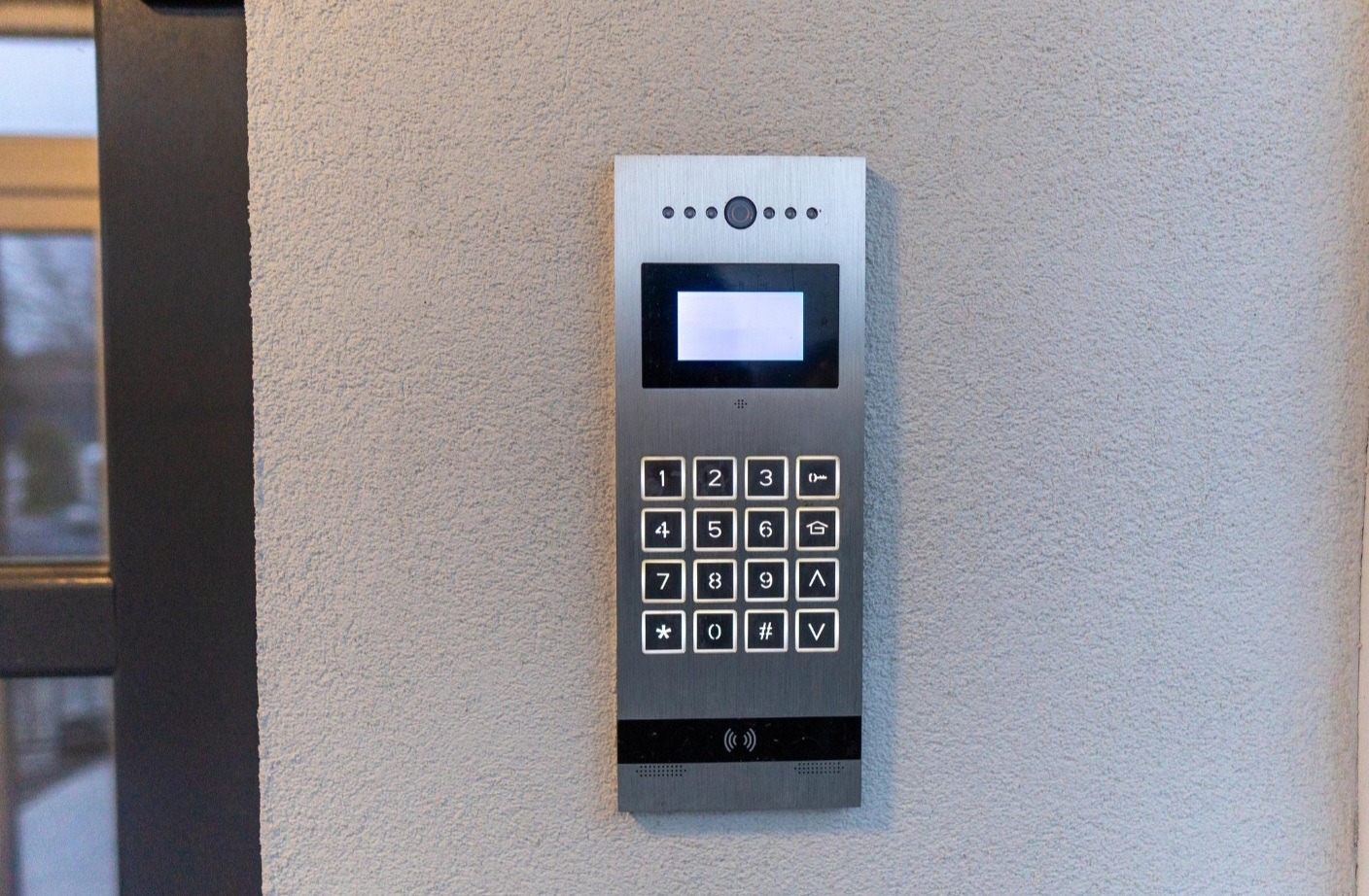 Nowoczesny panel domofonu z klawiaturą numeryczną i wyświetlaczem, zamontowany na teksturowanej ścianie zewnętrznej budynku. Widoczne przyciski z podświetleniem.