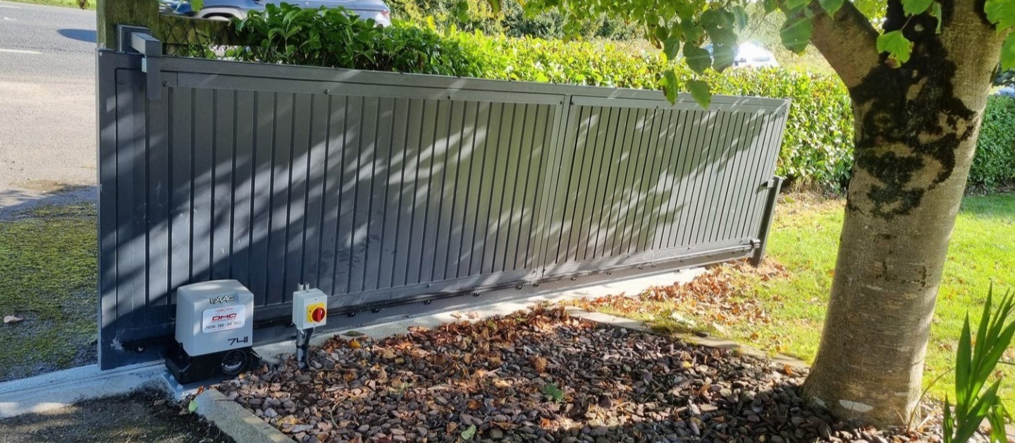 Automatyczna brama przesuwna z szarymi panelami pionowymi, widoczny napęd FAAC i skrzynka sterownicza, otoczenie zieleni i drzewa.