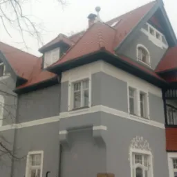 Szary dom z czerwonym dachem w stylu secesyjnym, widoczne detale architektoniczne i dekoracyjne elementy fasady, drzewo bez liści na pierwszym planie, pochmurna pogoda.