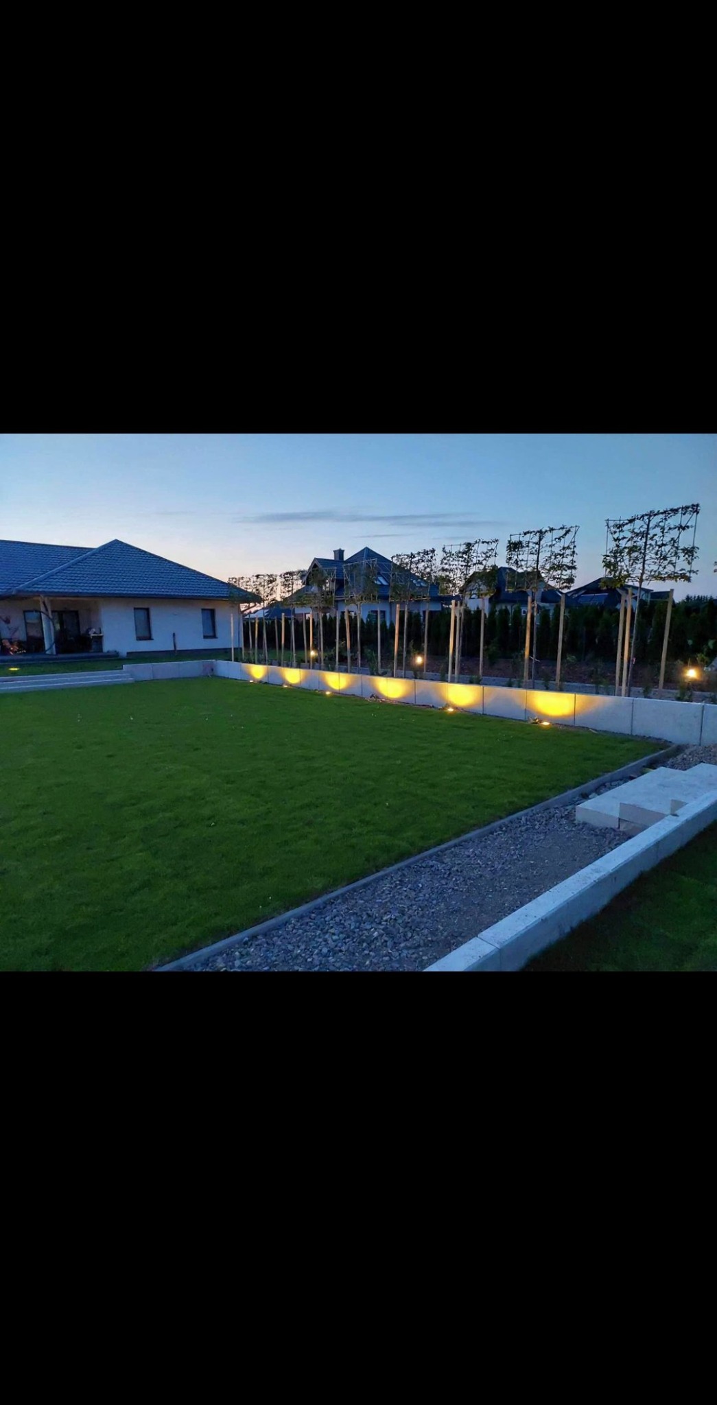 Zadbany trawnik z rolki oświetlony lampami wzdłuż murku. W tle dom z ogrodem i formowane drzewa. Widok o zmierzchu.