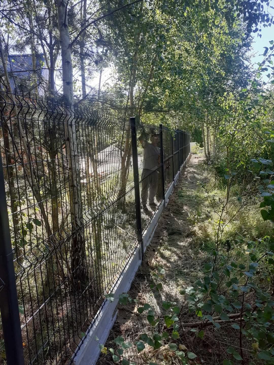 Długi, czarny panelowy płot na betonowej podmurówce, biegnący wzdłuż zarośniętej ścieżki, otoczony drzewami i krzewami. Widoczna osoba montująca ogrodzenie.