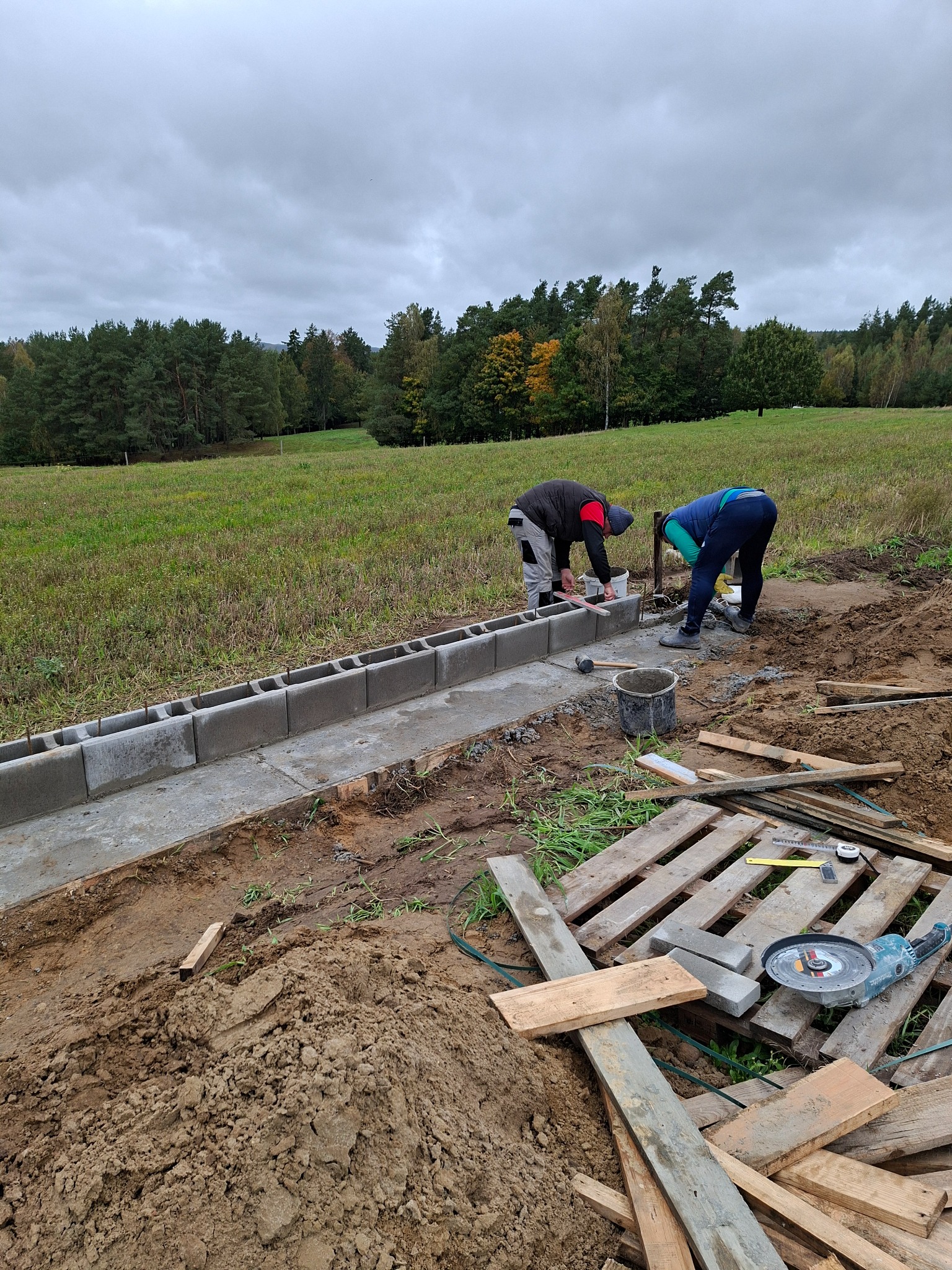 Układanie obrzeży z betonowych bloczków przez dwóch pracowników na tle łąki i lasu. Widoczne narzędzia i paleta z drewnem.