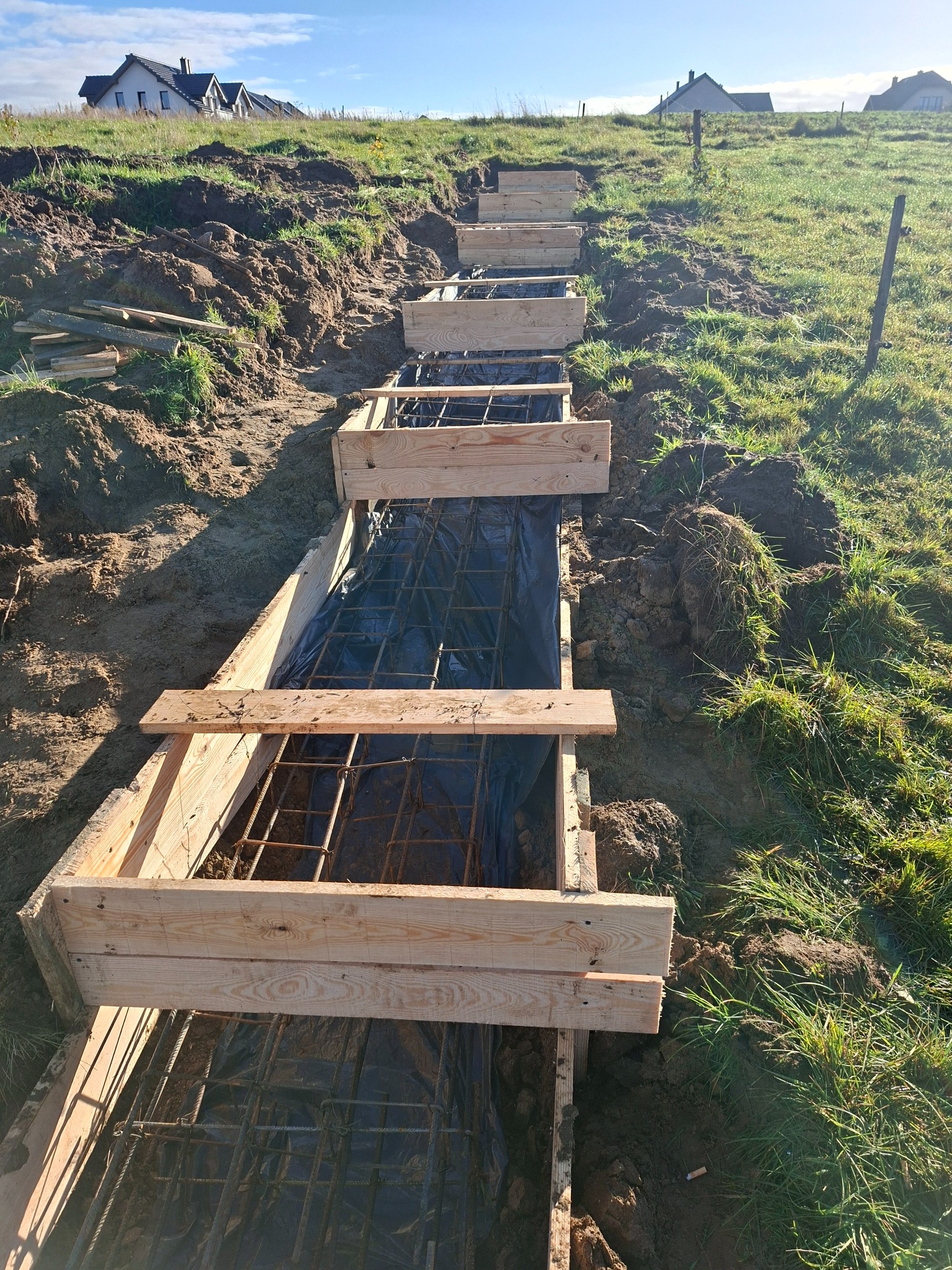Szkielet schodów z drewnianym szalunkiem i zbrojeniem stalowym na zboczu, zabezpieczony czarną folią. W tle domy i błękitne niebo.