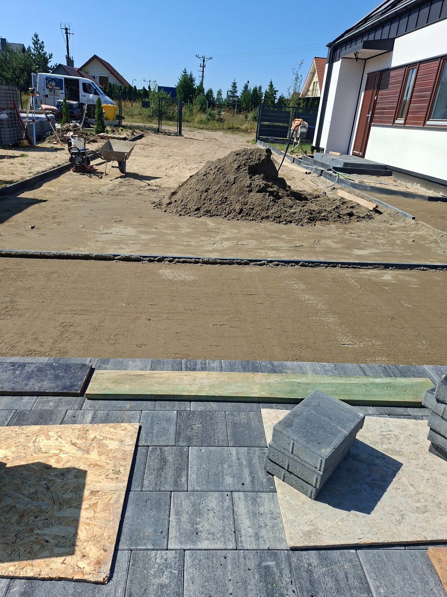 Etap układania kostki brukowej na posesji. Widoczny przygotowany teren, obrzeża, piasek, ułożona część kostki i paleta z kostką. W tle dom i sprzęt budowlany.