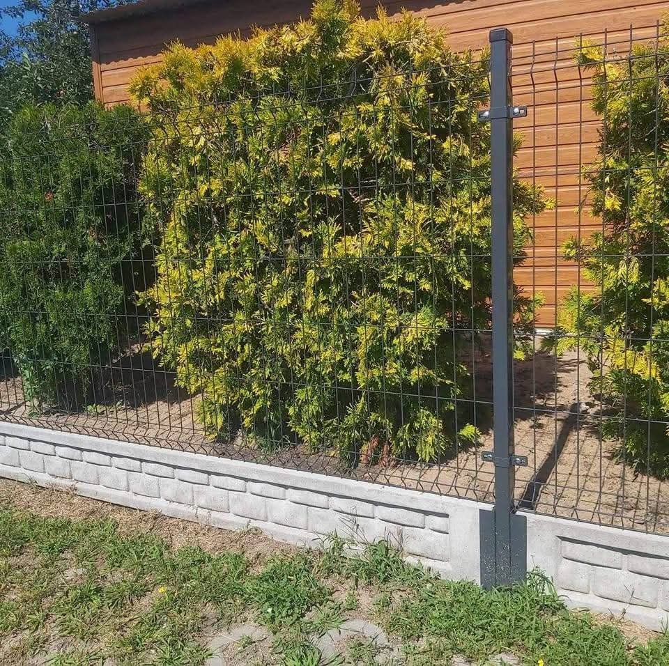 Nowoczesne ogrodzenie panelowe z podmurówką betonową i słupkami w kolorze grafitowym, na tle zielonych krzewów i drewnianego budynku.