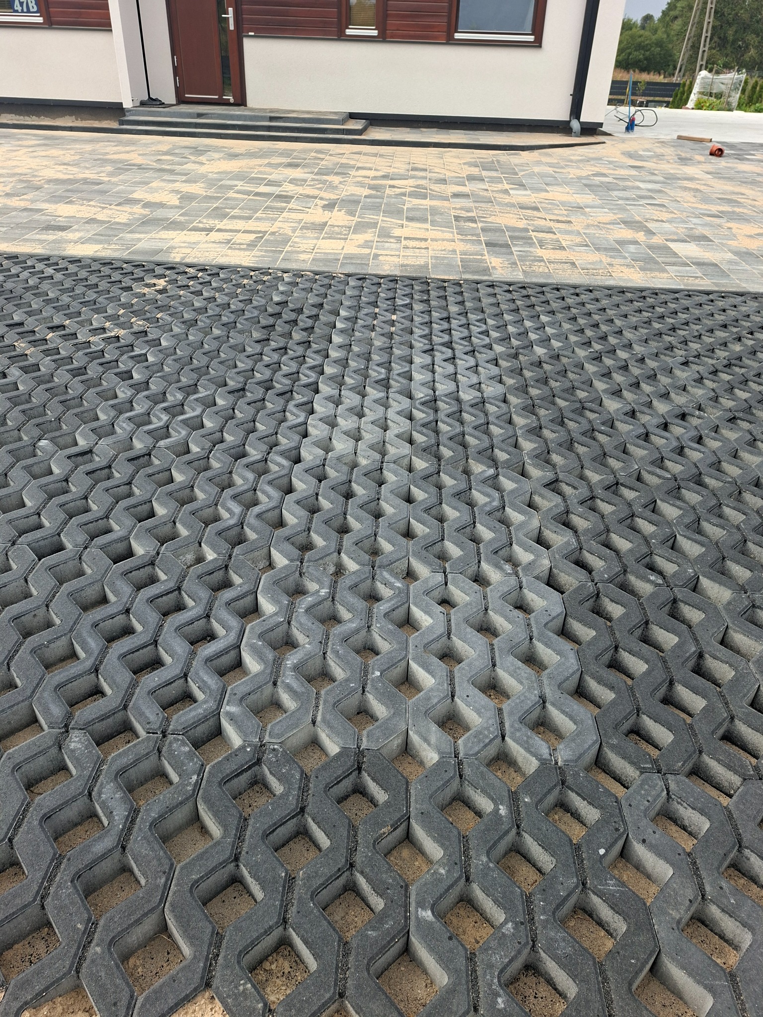 Ułożona szara ażurowa kostka brukowa na podjeździe przed domem, z widoczną częścią klasycznej kostki w tle. Widok z góry, podkreślający wzór i strukturę.