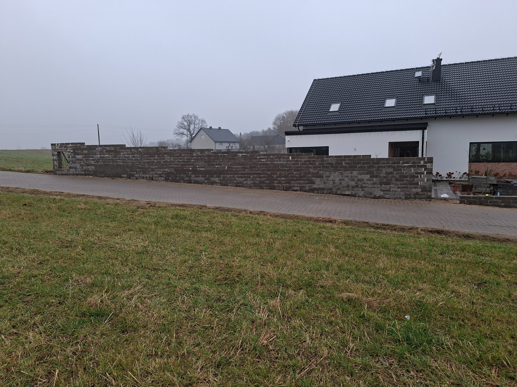Wznoszona z szarej kostki ogrodowa ściana oporowa na trawiastym terenie, z widocznym fragmentem domu z ciemnym dachem i kostką brukową. Pochmurne niebo w tle.