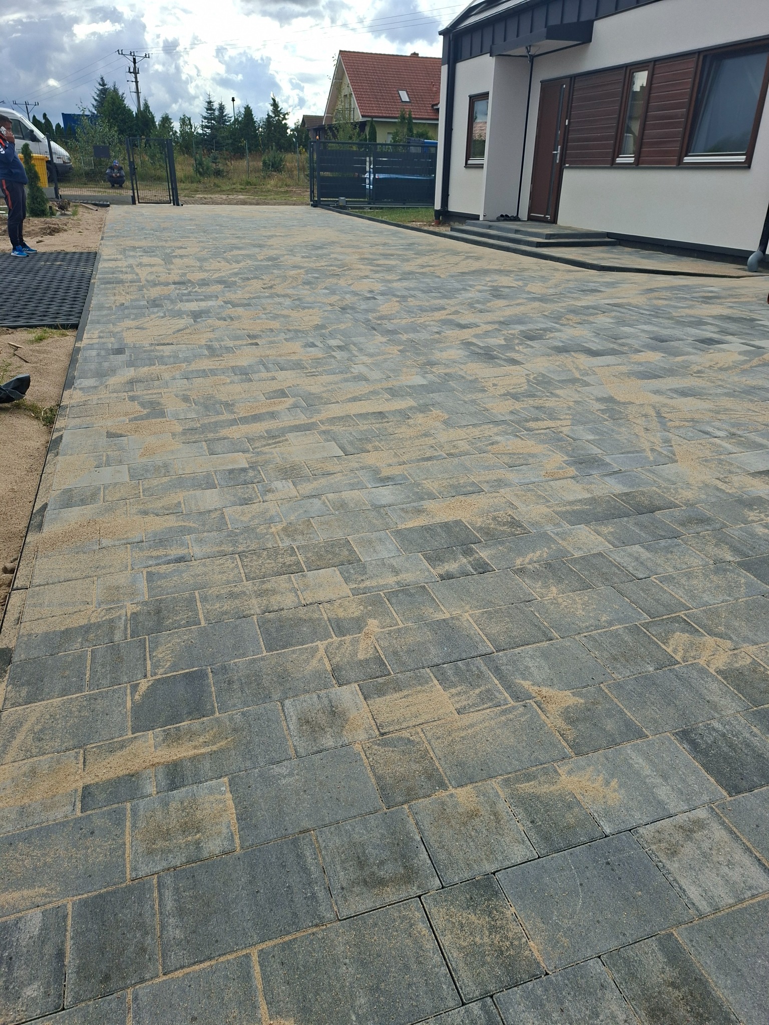 Ułożona, szara kostka brukowa na podjeździe, z piaskiem w szczelinach, prowadząca do nowoczesnego domu z brązowymi okiennicami. Widoczny fragment bramy w tle.