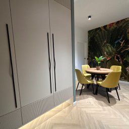 17 Studio Projektowe Sylwia Bierwiaczonek - Szara szafa na wymiar z frezowanymi detalami i długimi, czarnymi uchwytami. W tle okrągły stół z żółtymi krzesłami i tapeta w tropikalne wzory.