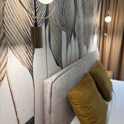 17 Studio Projektowe Sylwia Bierwiaczonek - Stylowa sypialnia z tapetą w motywy liści, lampką nocną z okrągłym kloszem i beżowym zagłówkiem. Złote poduszki na białej pościeli. Nowoczesne gniazdka elektryczne.