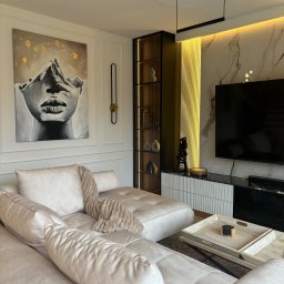 17 Studio Projektowe Sylwia Bierwiaczonek - Elegancki salon z narożnikiem w odcieniach beżu, obrazem z motywem górskim i złotymi akcentami, podświetlaną ścianą z marmuru i nowoczesnym oświetleniem.