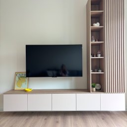 17 Studio Projektowe Sylwia Bierwiaczonek - Nowoczesna szafka RTV z półkami i lamelami w odcieniach drewna, wisząca nad podłogą. Na blacie dekoracje: plakat Capri i żółta miseczka. Minimalistyczny design.