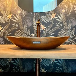 17 Studio Projektowe Sylwia Bierwiaczonek - Miedziana umywalka w kształcie łodzi na drewnianym blacie, z baterią w kolorze miedzi. W tle lustro i ściana w tropikalne wzory. Nowoczesna aranżacja łazienki.