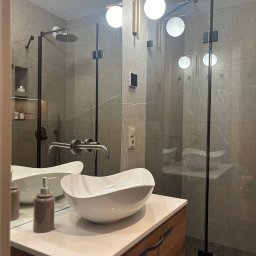 17 Studio Projektowe Sylwia Bierwiaczonek - Nowoczesna łazienka z szarą glazurą, kabiną prysznicową typu walk-in i umywalką nablatową. Uwagę zwraca drewniana szafka pod umywalką i designerskie oświetlenie.