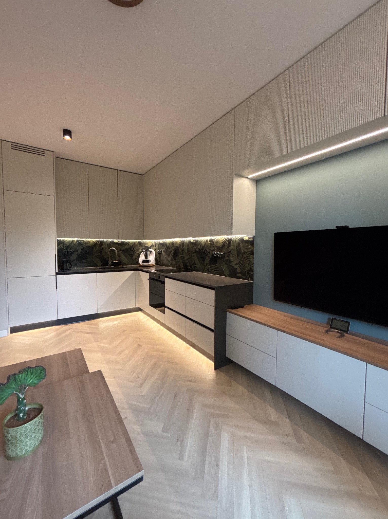Nowoczesna kuchnia z szafkami w odcieniach szarości, podświetleniem LED i blatem z motywem roślinnym. Parkiet w jodełkę i stół z rośliną doniczkową.