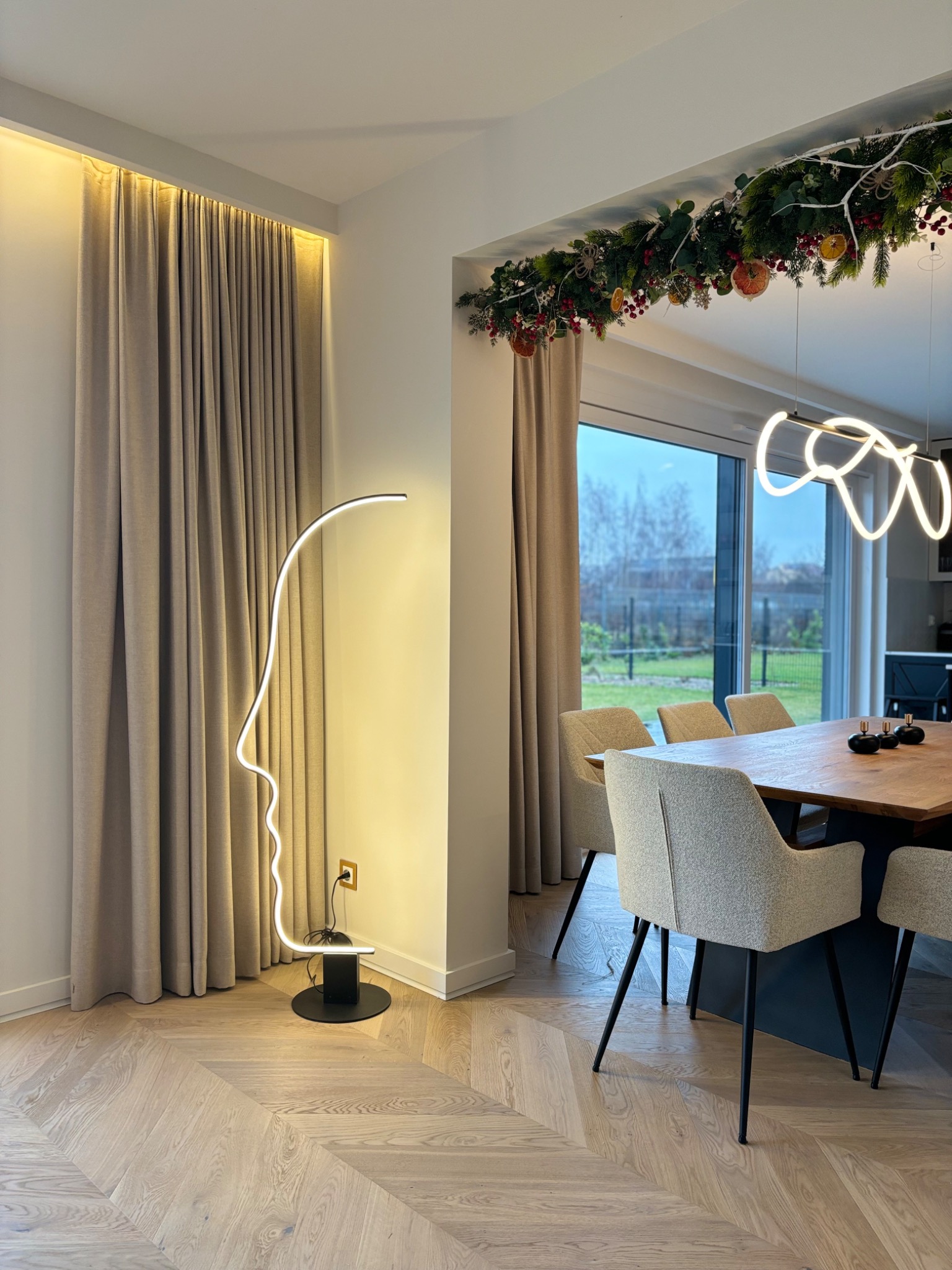 Nowoczesna jadalnia z parkietem w jodełkę, beżowymi zasłonami z podświetleniem LED i lampą podłogową w kształcie profilu twarzy. Świąteczny wystrój nad oknem.