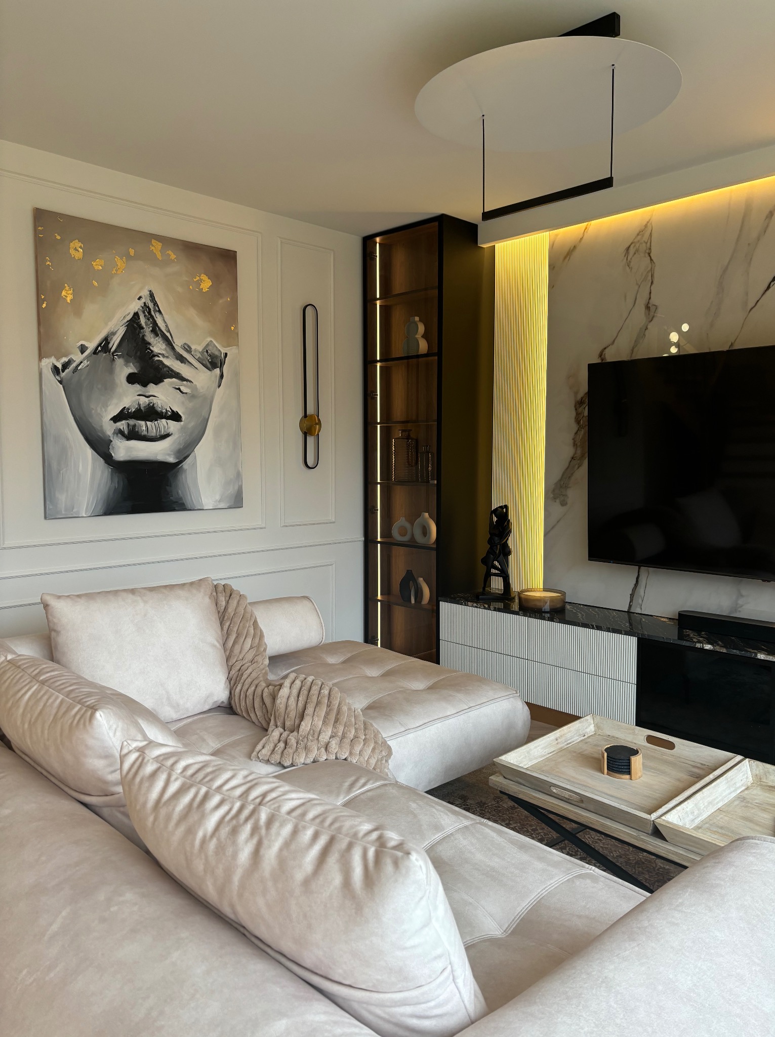 Elegancki salon z narożnikiem w odcieniach beżu, obrazem z motywem górskim i złotymi akcentami, podświetlaną ścianą z marmuru i nowoczesnym oświetleniem.