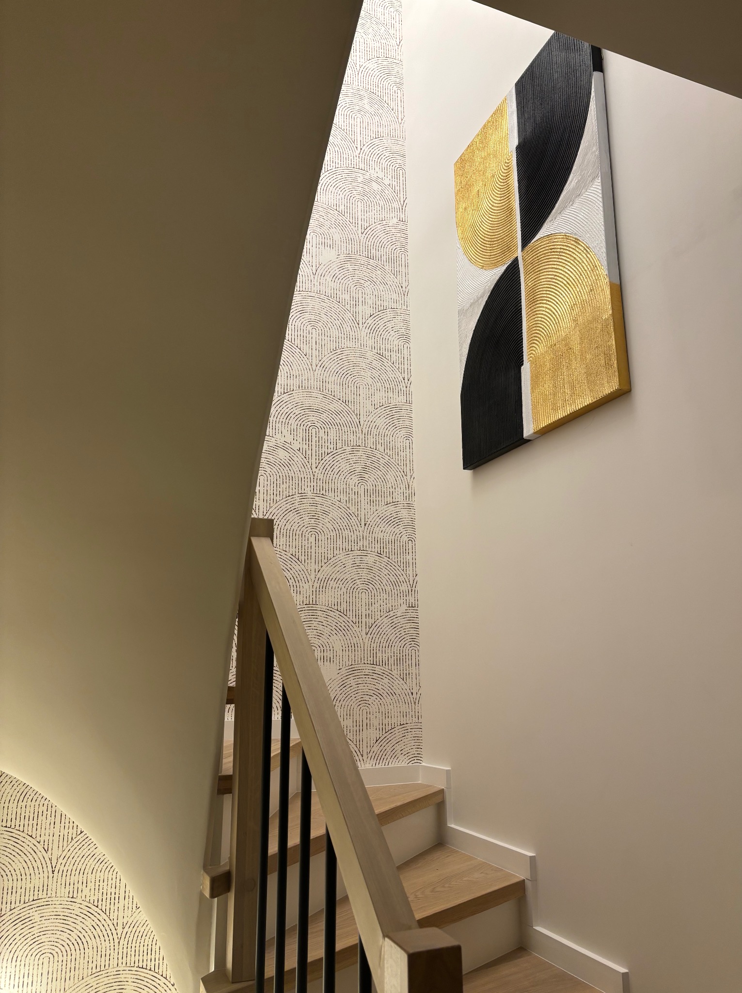 Schody z drewnianymi stopniami i czarną balustradą, ściana z wzorzystą tapetą i obrazem w geometryczne wzory w odcieniach złota, czerni i bieli. Nowoczesne wykończenie.