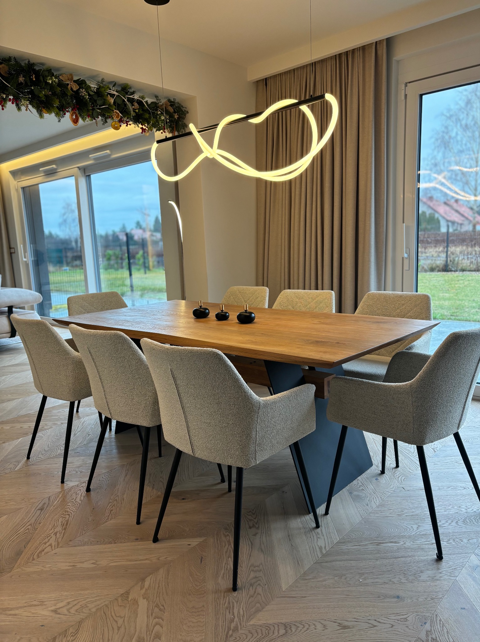 Drewniany stół z krzesłami w jadalni w Księginicach, oświetlony nowoczesną lampą. Parkiet w jodełkę, duże okna z widokiem na ogród. Dekoracje świąteczne.
