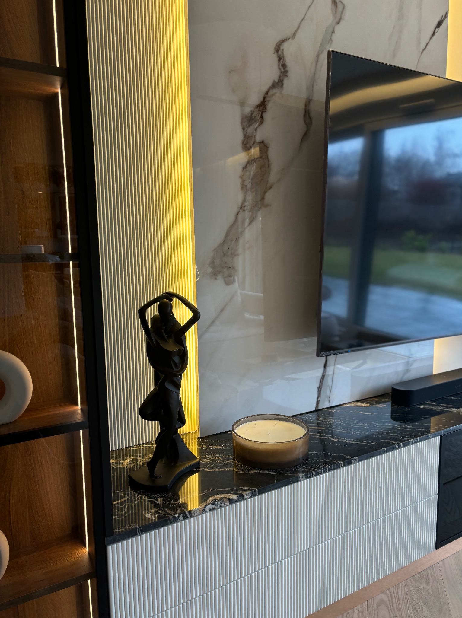 Nowoczesna aranżacja salonu z marmurową ścianą TV, podświetlaną szafką RTV z czarnym blatem i dekoracyjną figurką. Widoczna część drewnianej witryny z oświetleniem.