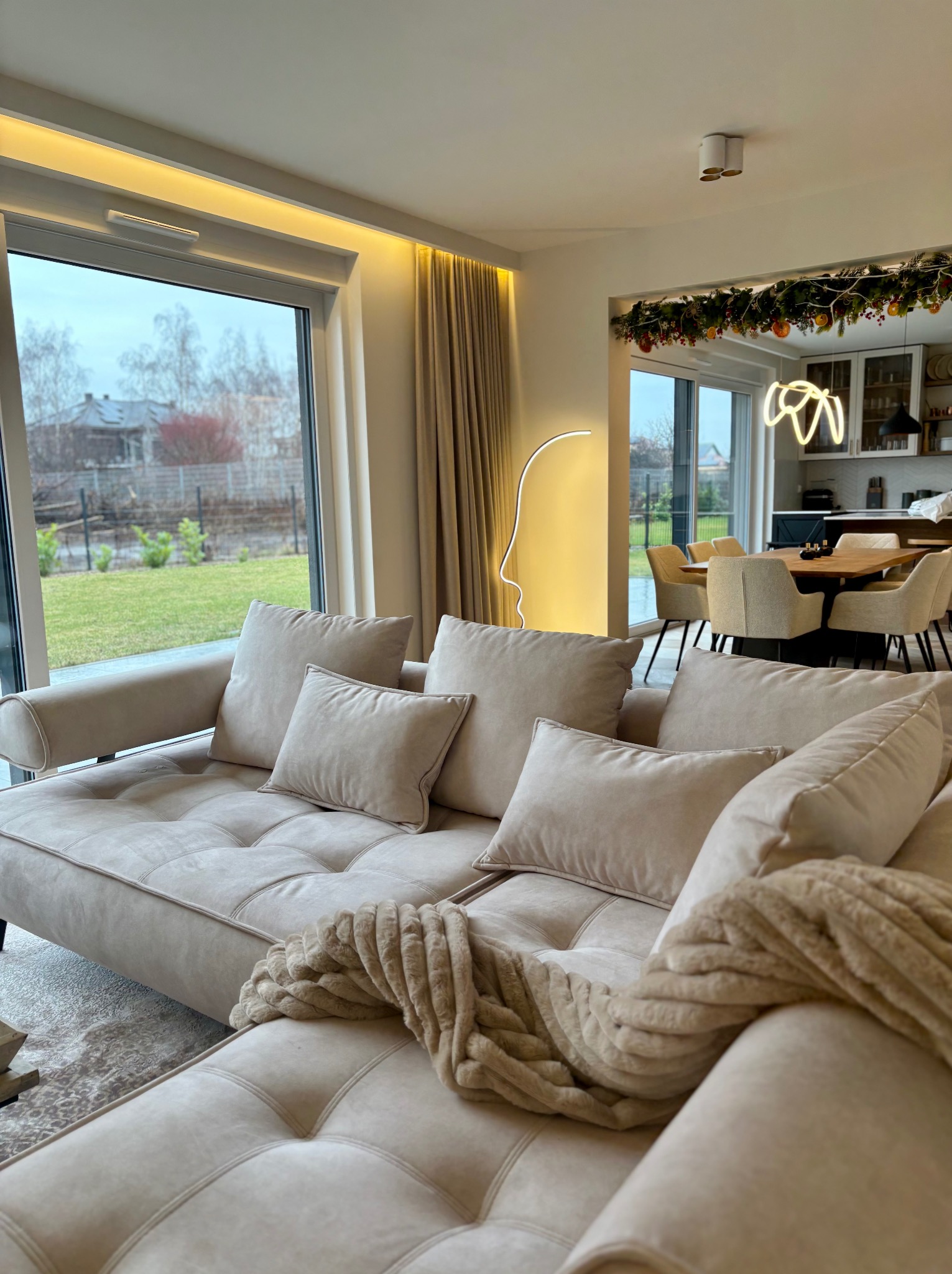 Jasny salon z beżową sofą, poduszkami i kocem. W tle widoczny stół z krzesłami i dekoracje świąteczne. Duże okno z widokiem na ogród.