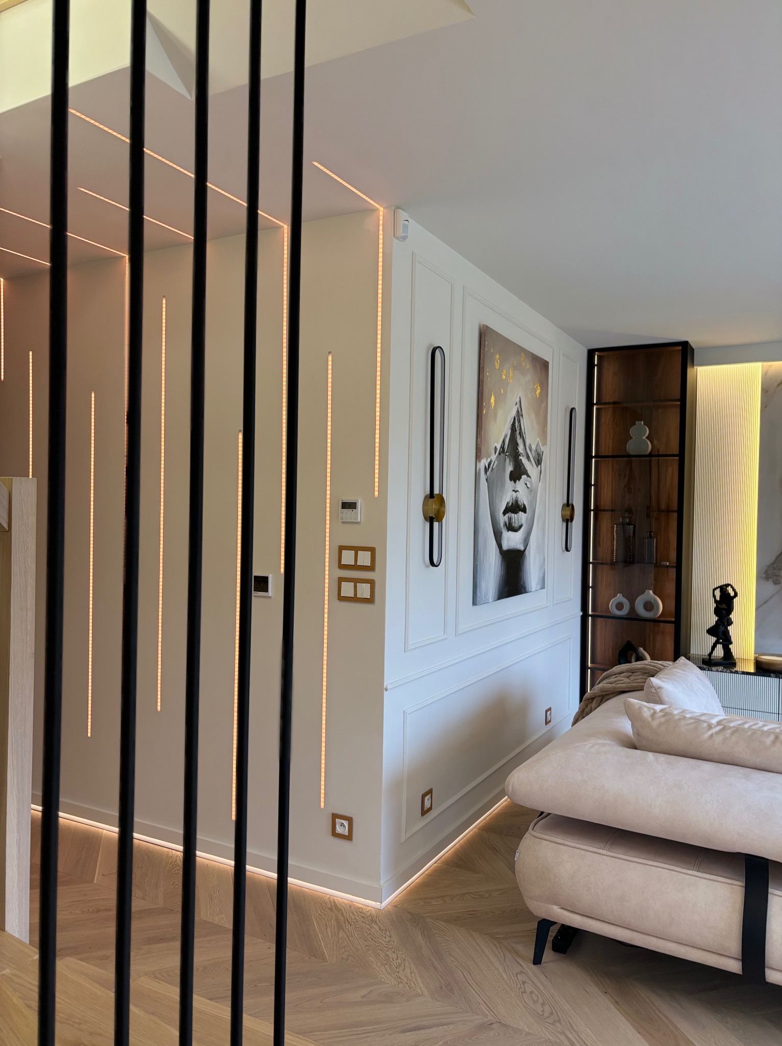 Nowoczesne wnętrze: parkiet w jodełkę, ściana z dekoracyjnymi listwami, oświetlenie LED, obraz i regał z podświetleniem. Widok zza czarnych, pionowych elementów.