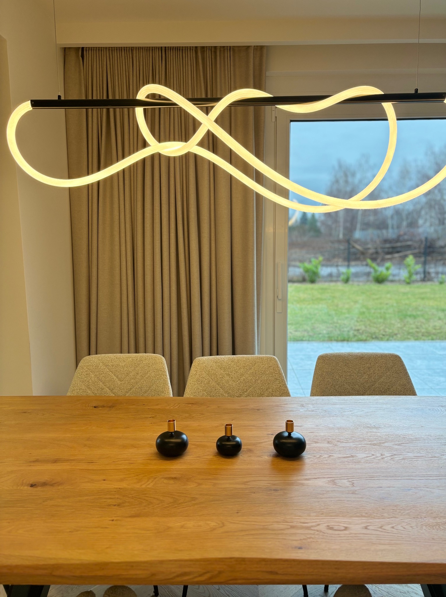 Stół z drewna dębowego, trzy czarne wazoniki, nowoczesna lampa LED o fantazyjnym kształcie, krzesła w tle, widok na ogród przez okno.
