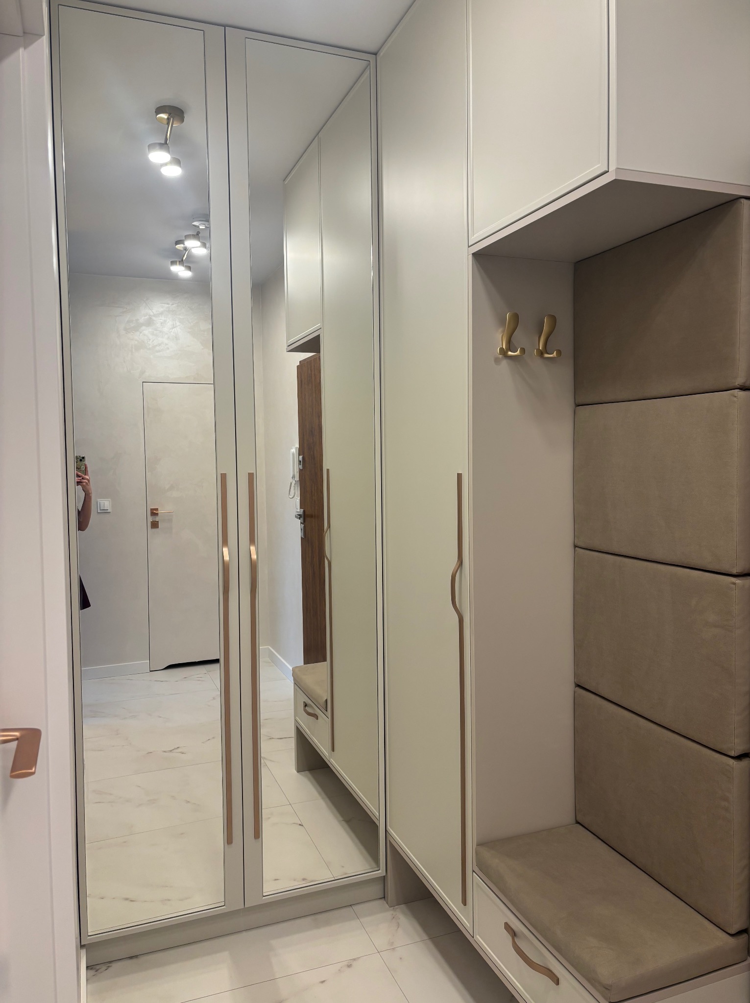 Jasna szafa wnękowa z lustrami i siedziskiem w przedpokoju. Minimalistyczny design, złote akcenty, odbicie wnętrza mieszkania. Nowoczesne wykończenie.