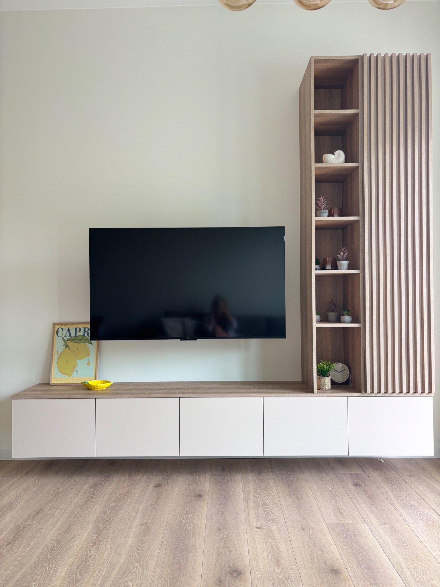 Nowoczesna szafka RTV z półkami i lamelami w odcieniach drewna, wisząca nad podłogą. Na blacie dekoracje: plakat Capri i żółta miseczka. Minimalistyczny design.