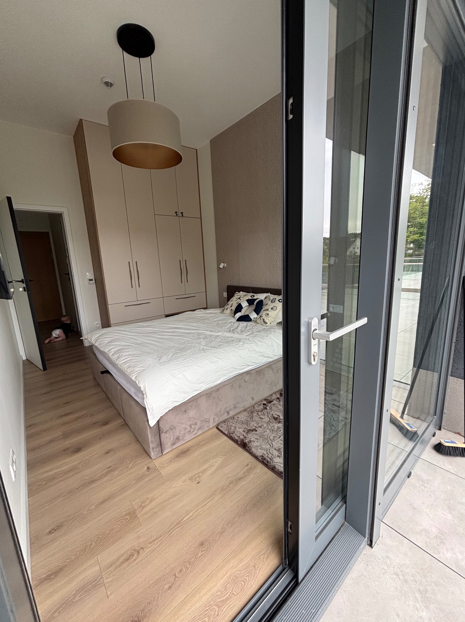 Jasna sypialnia z dużym łóżkiem, szafą w zabudowie i wyjściem na balkon przez przesuwne drzwi. Widoczny parkiet i dywanik. W tle, w drzwiach, małe dziecko.