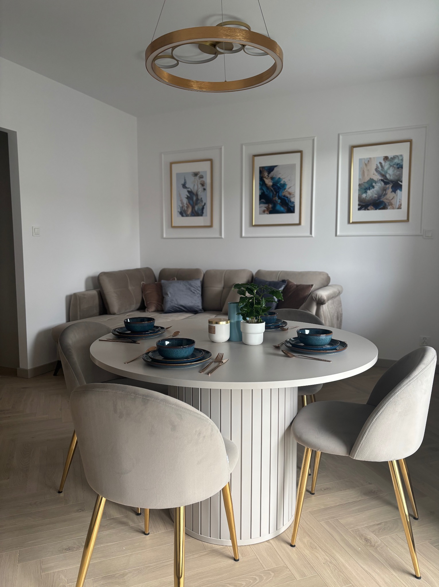 Jadalnia w neutralnych barwach: okrągły stół z nakryciem, welurowe krzesła na złotych nogach, sofa i obrazy w tle. Nowoczesny design z dbałością o detale.