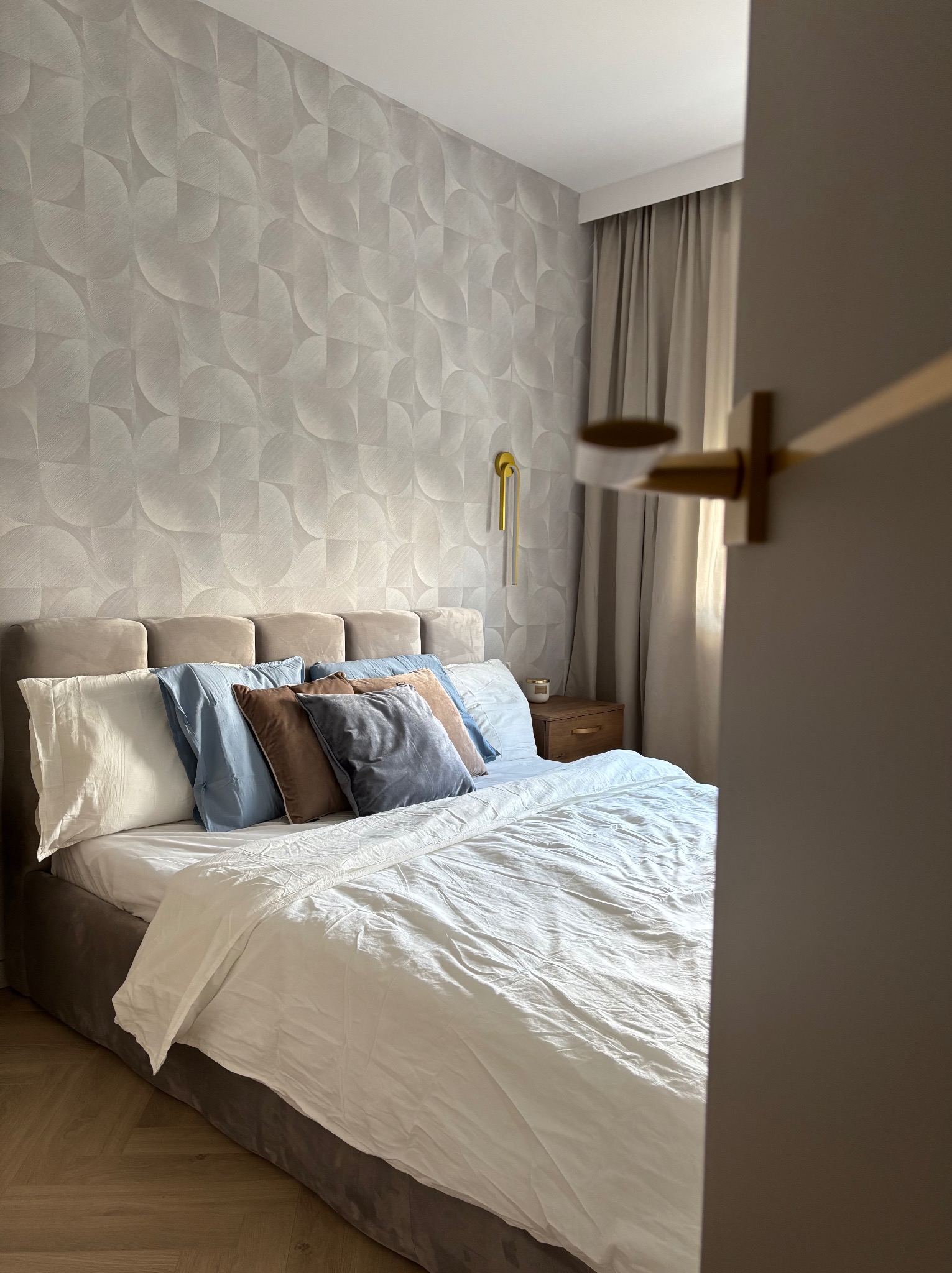 Nowoczesna sypialnia z łóżkiem w odcieniach beżu i szarości, wzorzysta tapeta i drewniana podłoga. Widok zza uchylonych drzwi.