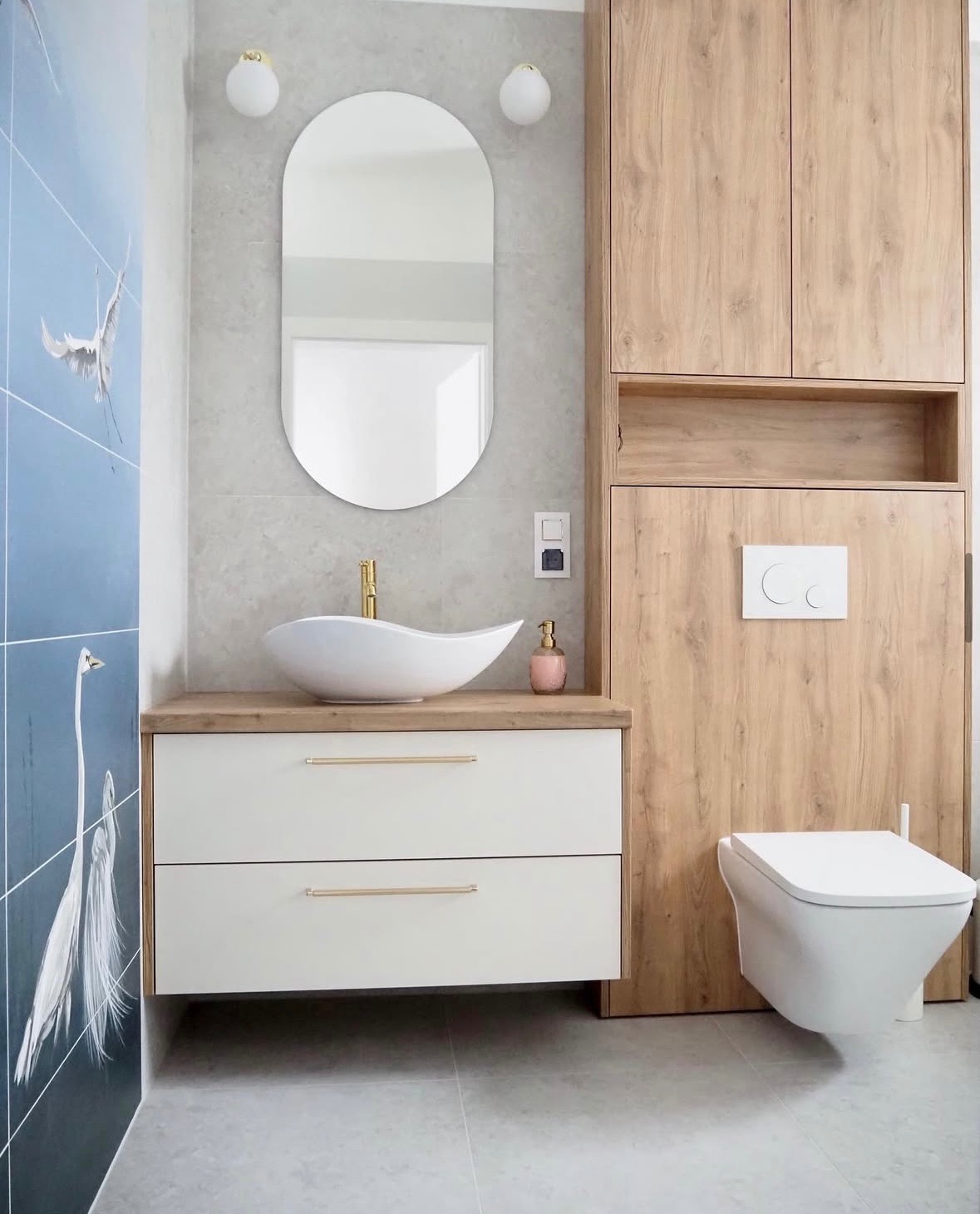 Nowoczesna łazienka z umywalką nablatową, lustrem w kształcie pigułki i szafką z drewnianym dekorem. Na ścianie płytki z motywem żurawi.