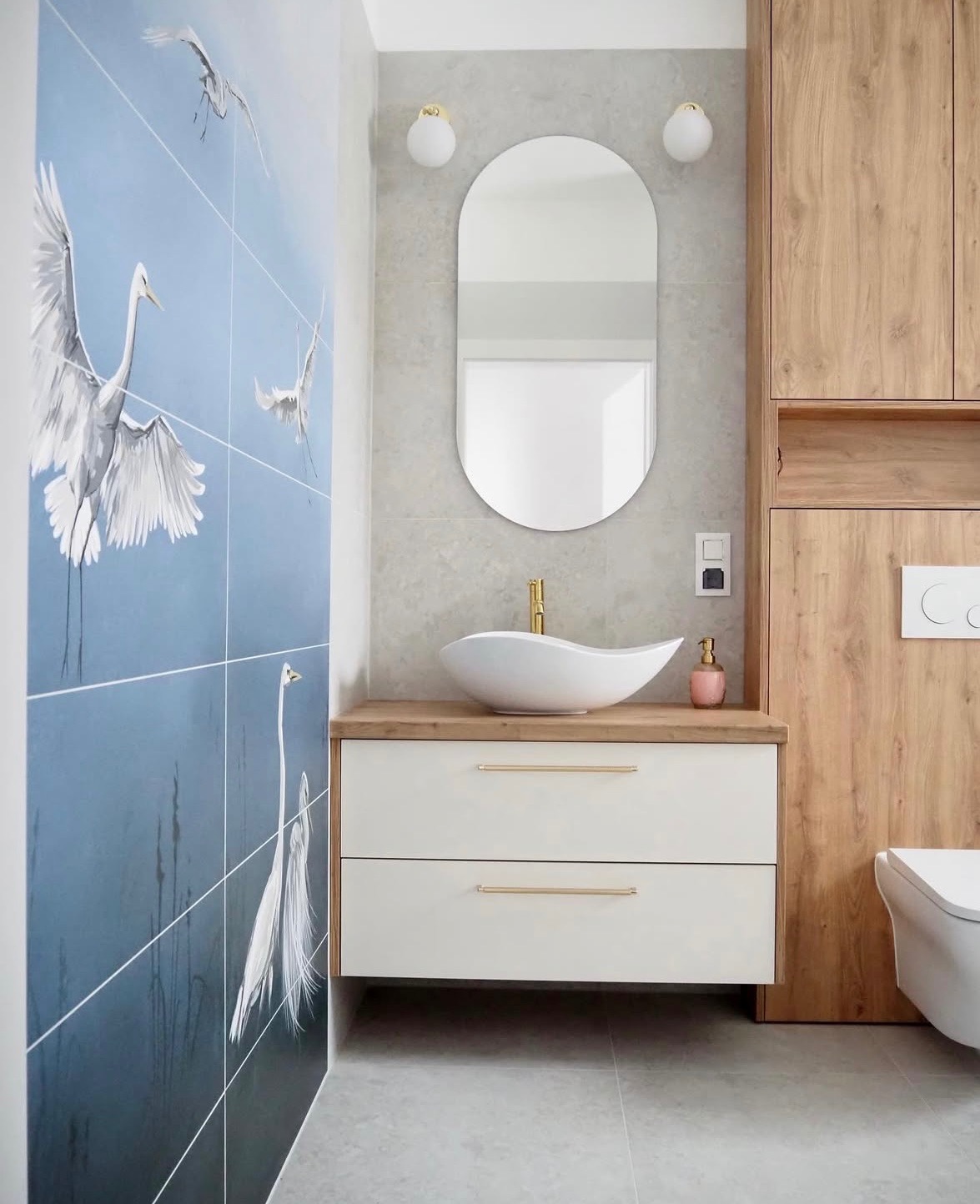 Nowoczesna łazienka z umywalką nablatową, lustrem w kształcie migdała i płytkami z motywem żurawi. Drewniane szafki i złote akcenty dodają elegancji.