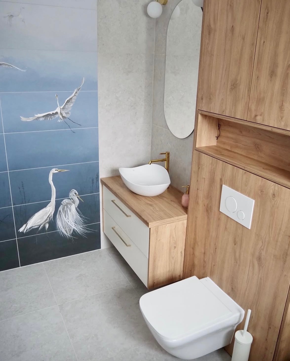 Nowoczesna łazienka z umywalką nablatową, drewnianą szafką i płytkami z motywem ptaków. Minimalistyczne oświetlenie i owalne lustro dopełniają aranżację.