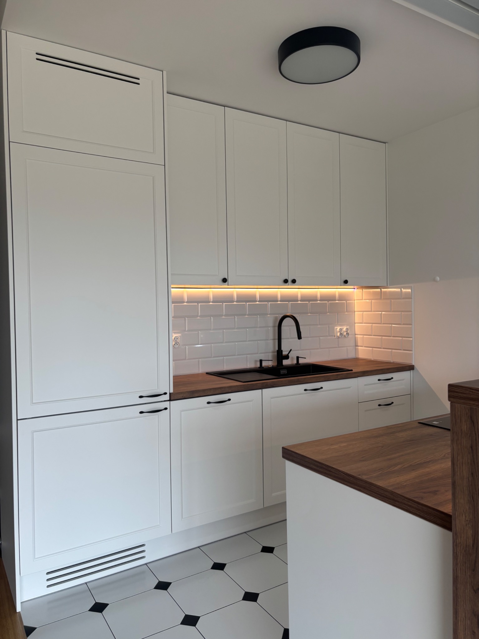 Biała kuchnia z drewnianym blatem i czarną armaturą. Uwagę przykuwają płytki w stylu retro na podłodze i podświetlenie LED nad blatem.