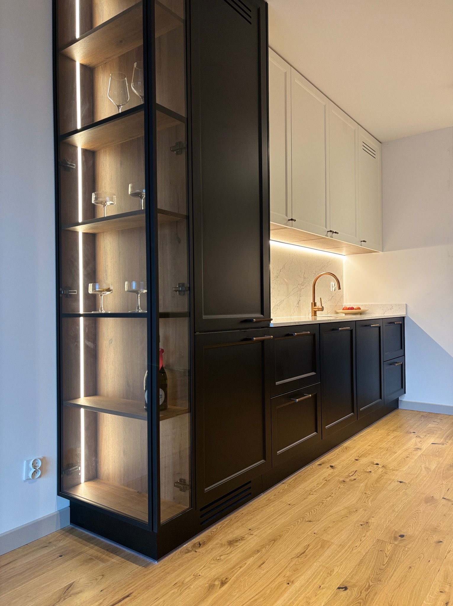Nowoczesna kuchnia z czarnymi szafkami dolnymi i białymi górnymi, drewnianym blatem i podświetlaną witryną z kieliszkami. Minimalistyczny design, ciepłe oświetlenie.