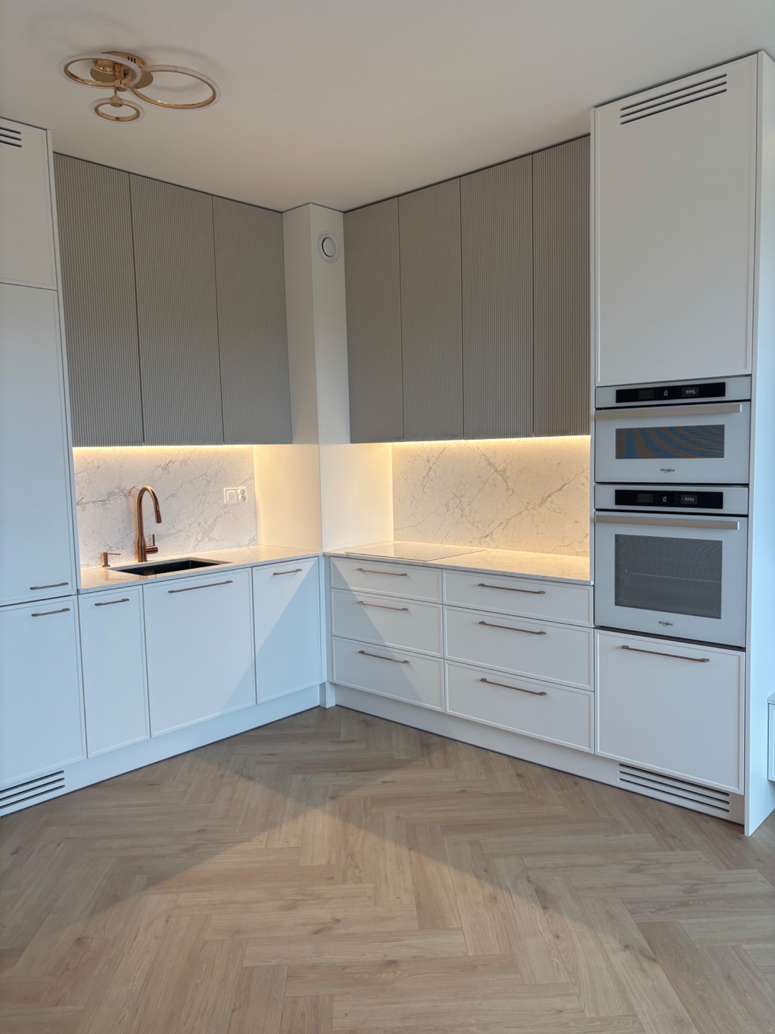 Nowoczesna kuchnia z białymi szafkami, marmurowym blatem i złotą armaturą. Górne szafki w odcieniach szarości z subtelnym, pionowym frezowaniem. Parkiet w jodełkę.